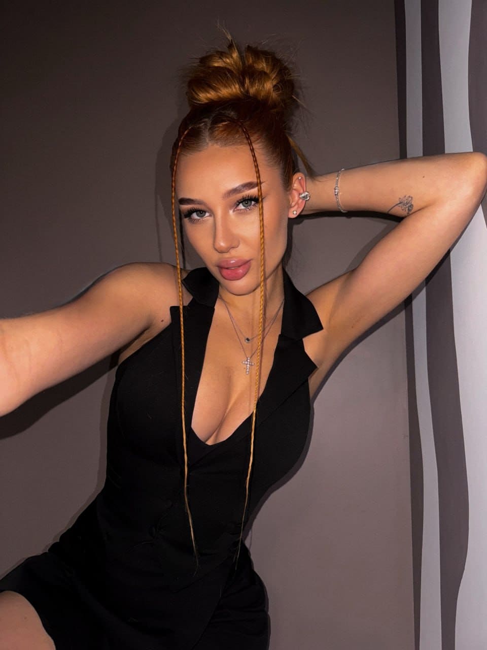 With hair in a high bun, she poses with arm raised behind head in a black plunging neckline top against striped walls.