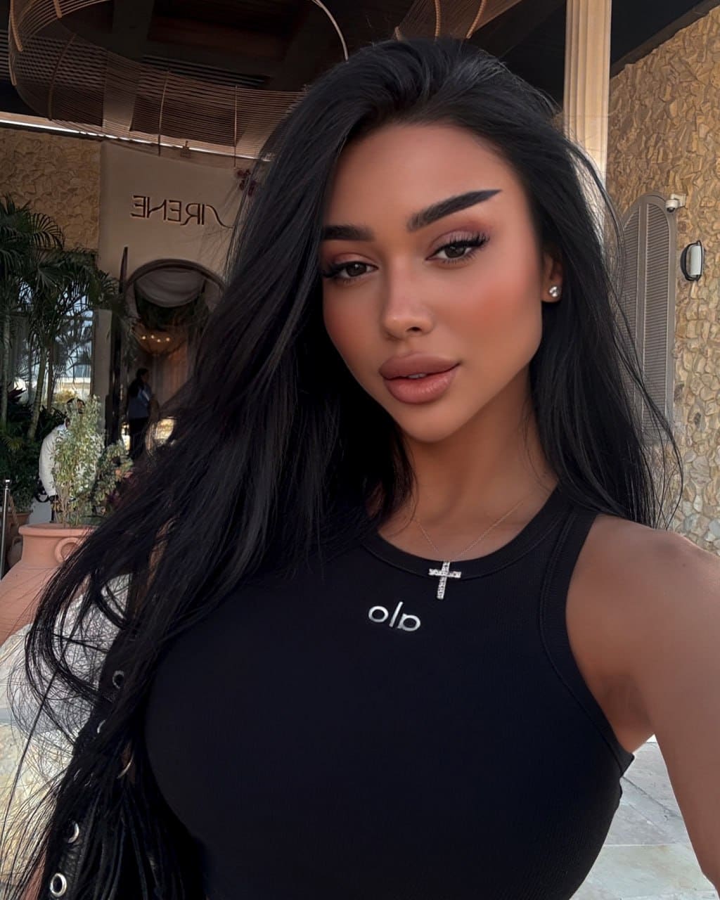 Inara takes a selfie outdoors against stone walls and plants, wearing a black sleeveless top with a logo and a cross necklace.
