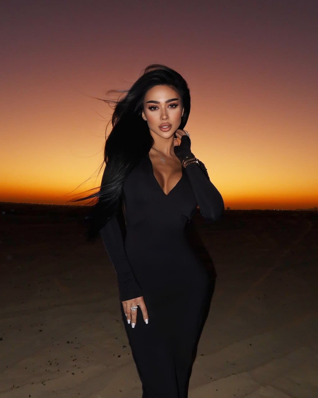 Inara stands in a desert at sunset, posing with a hand near her neck in a floor-length black dress with long sleeves and deep V-neck.