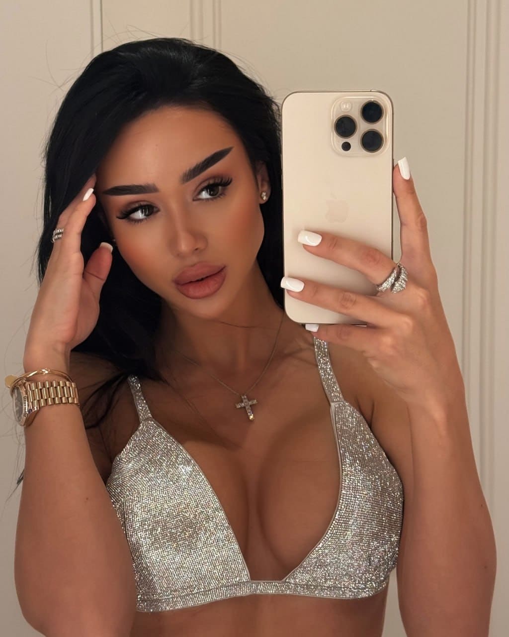 A mirror selfie shows Inara in a silver sequined bra top, gold watch, and cross necklace, one hand near her head holding a phone.