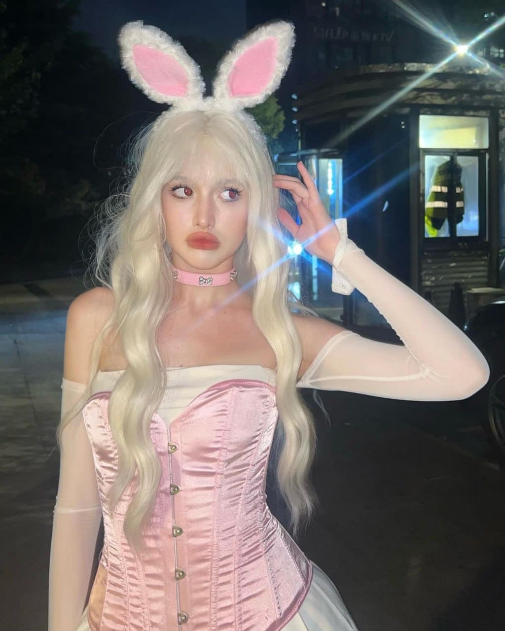 At night, she poses outdoors in a pink satin corset and bunny ears, raising one hand near her head.
