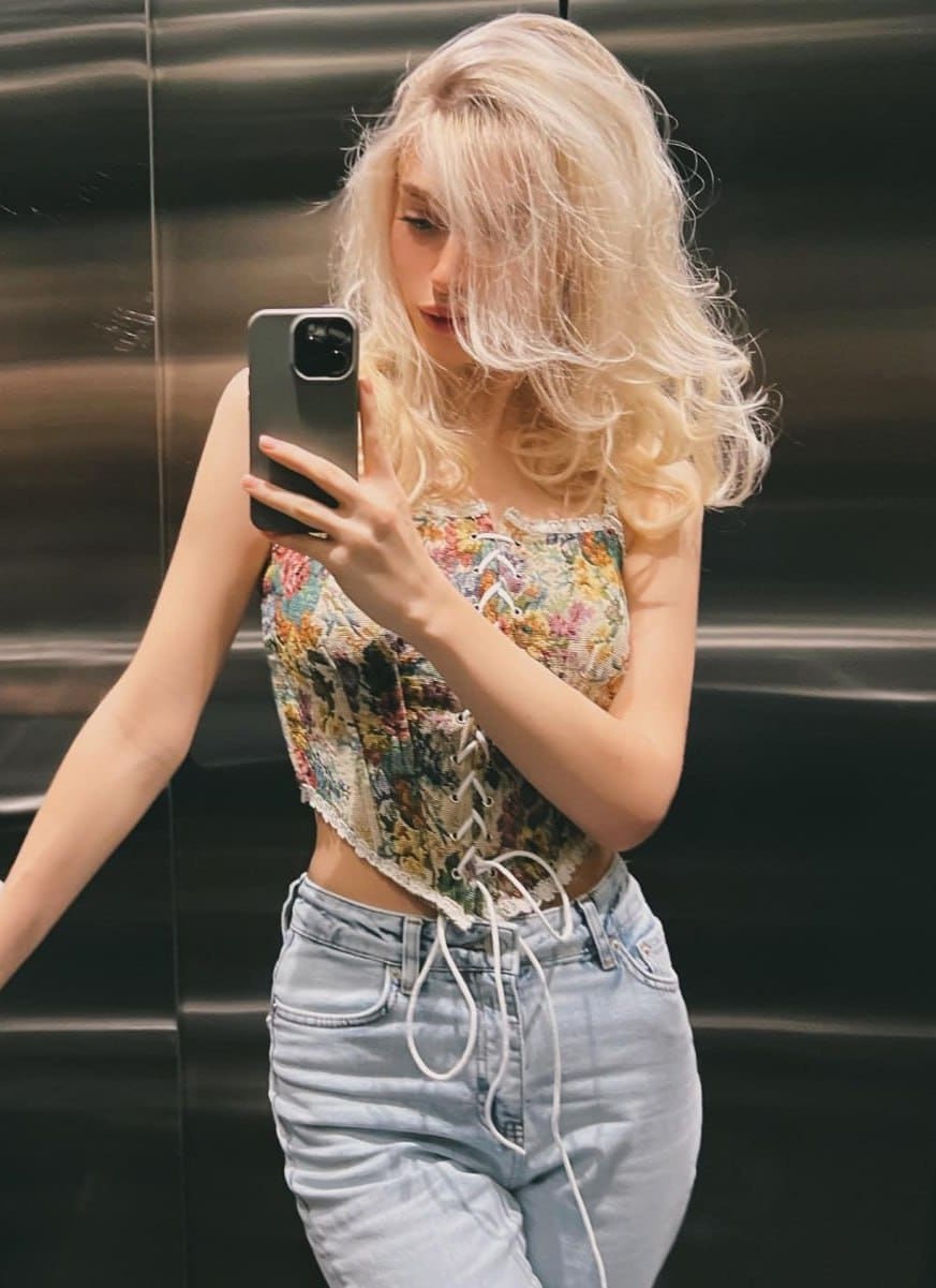 A mirror selfie in an elevator shows Laura in a floral lace-up corset top and light blue jeans.