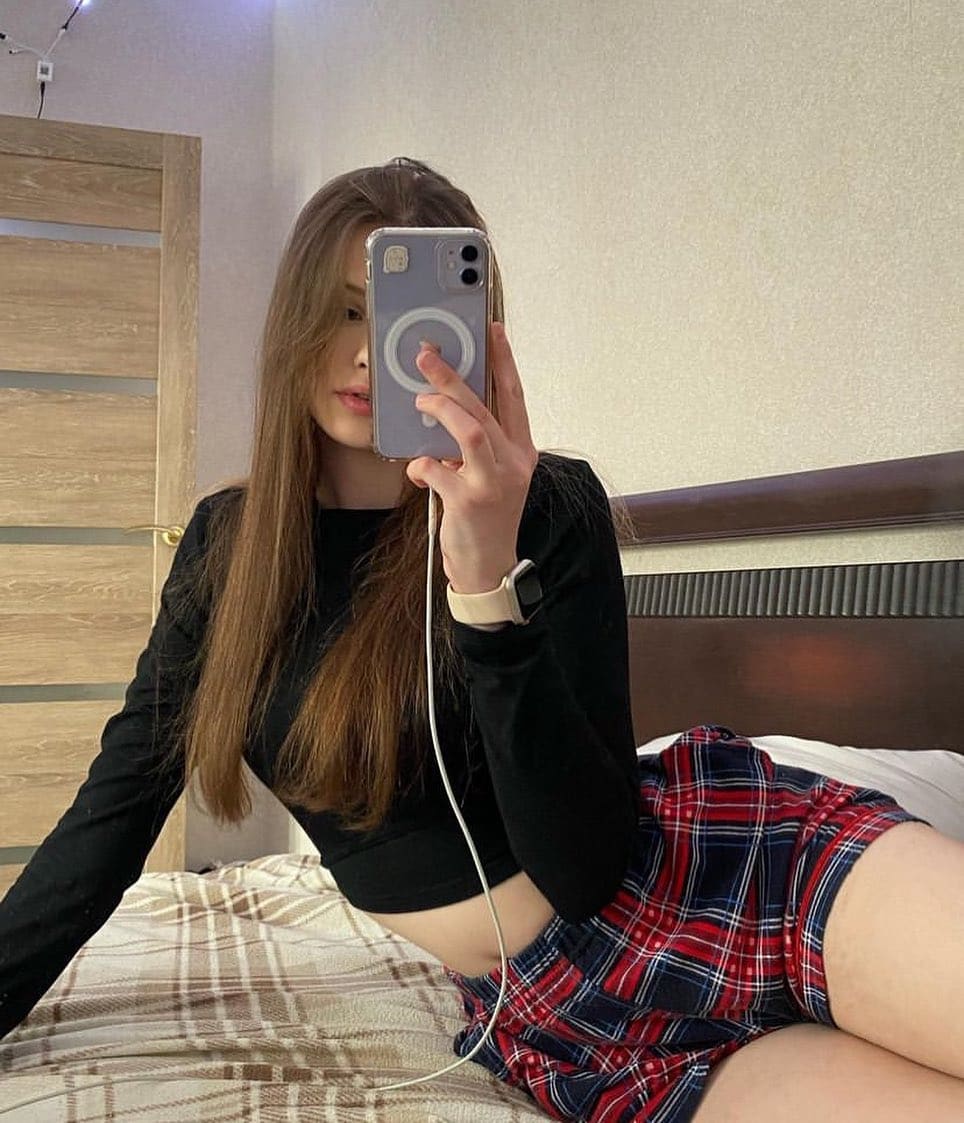 Sitting on a bed, Ava takes a mirror selfie wearing a black crop top and red plaid mini skirt.