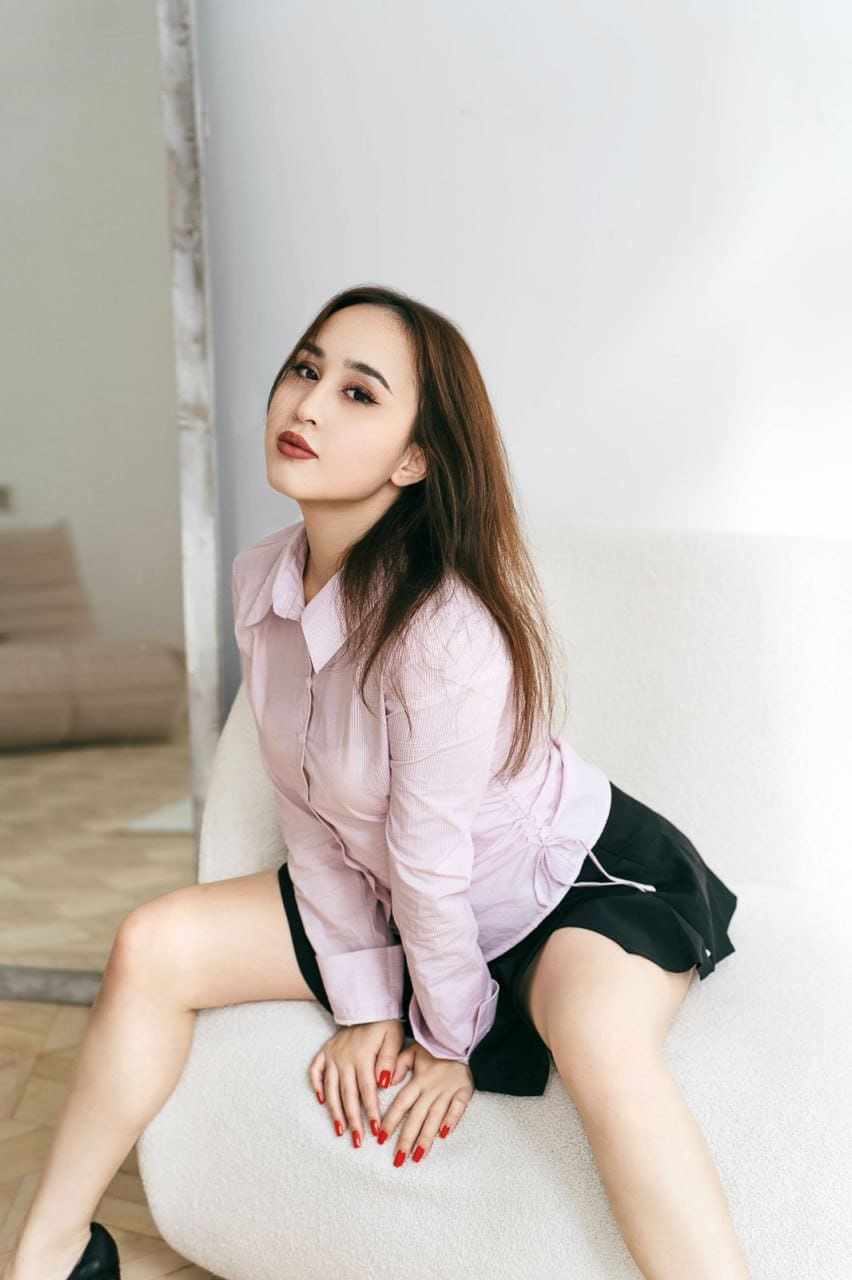 Sonya sits on a white chair in a pink shirt and black skirt, legs spread with hands resting between her thighs.