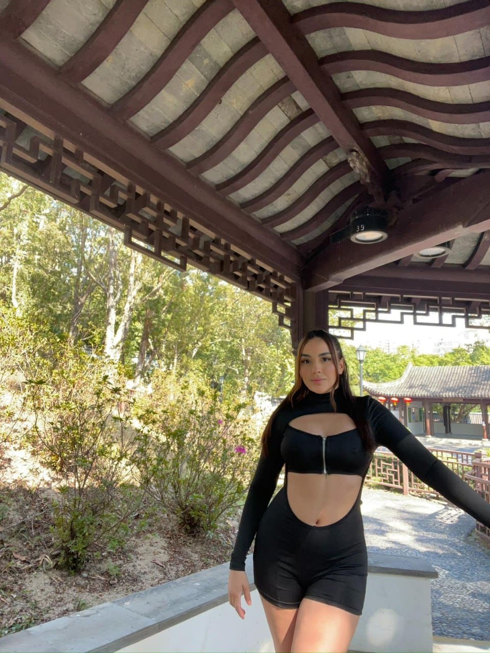 Under a wooden pergola, she extends one arm while wearing a black zippered bodysuit with midriff cutouts.