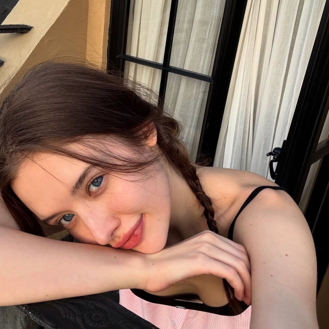 She rests her chin on crossed arms, wearing a pink and white striped top against sheer white curtains.