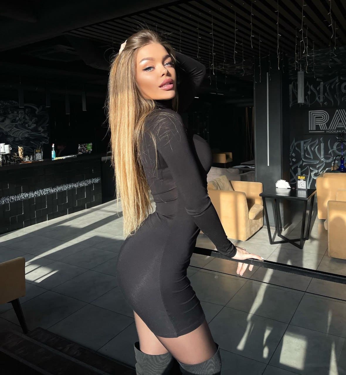 Standing in profile in a lounge, Halia wears a tight black mini dress and thigh-high boots, resting one hand on her head.