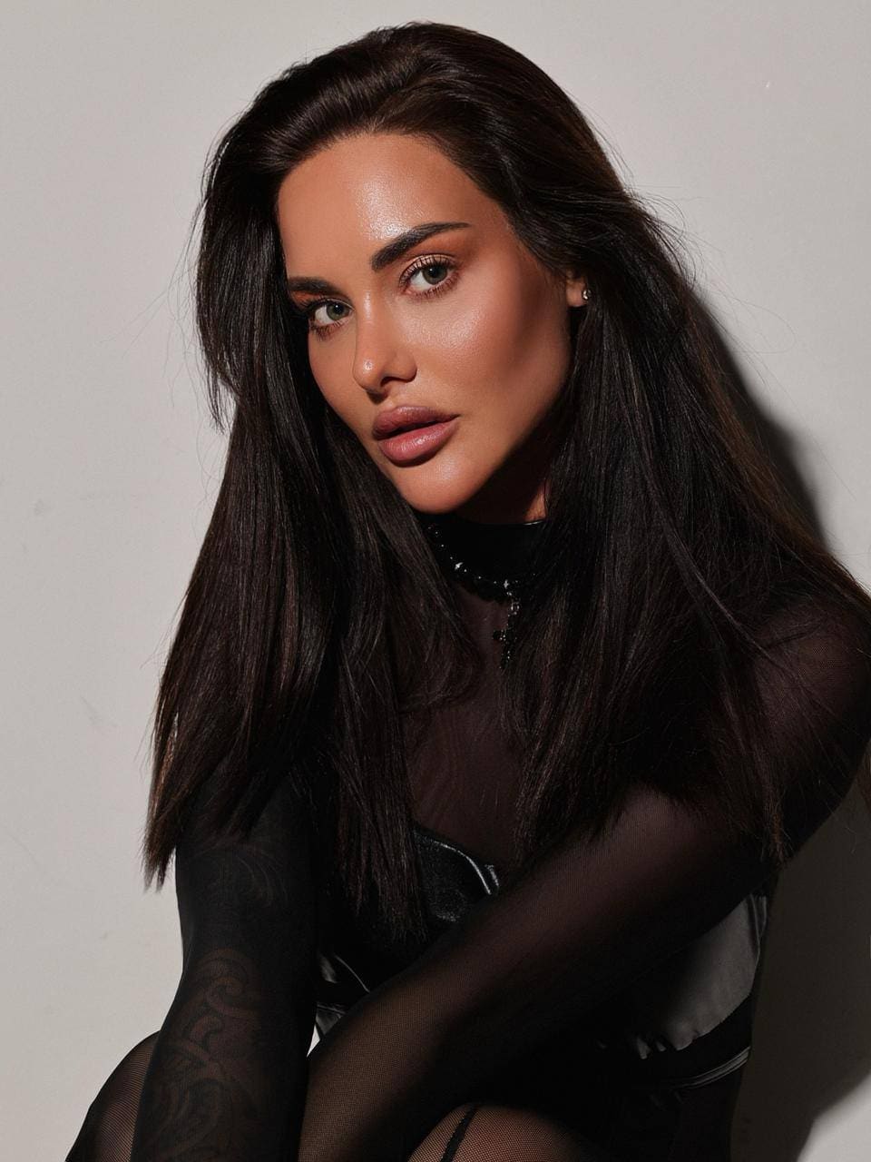 Eliana poses with arms crossed against a plain background, wearing a sheer black mesh top over dark undergarments.