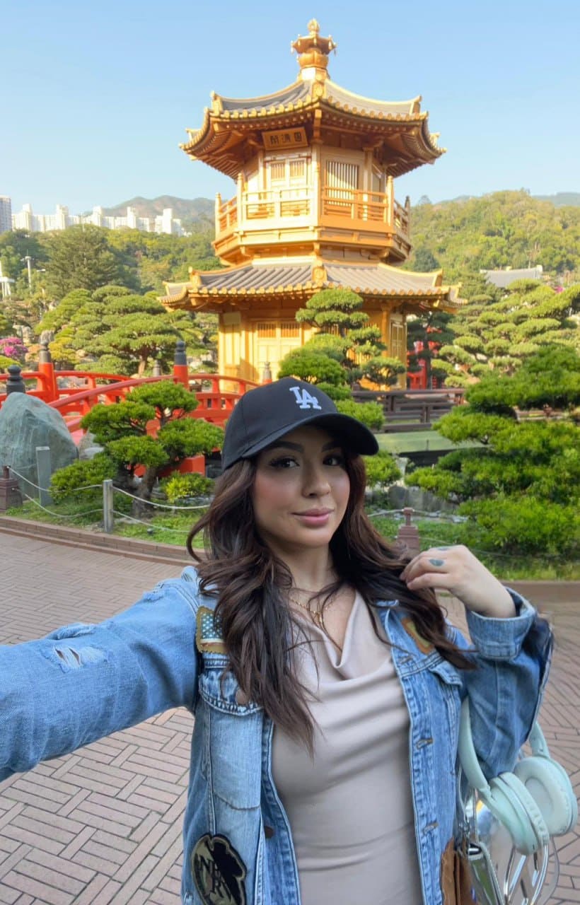 Rosa wears a black cap and denim jacket, holding mint headphones outdoors near a golden pagoda and red bridge.