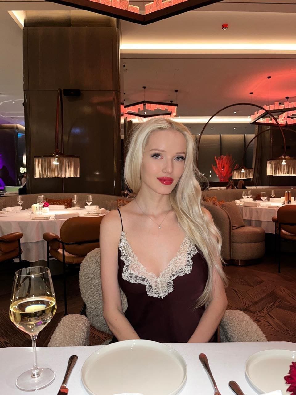 Nova sits at a restaurant table in a dark maroon slip dress with white lace trim, a glass of white wine visible in the foreground.