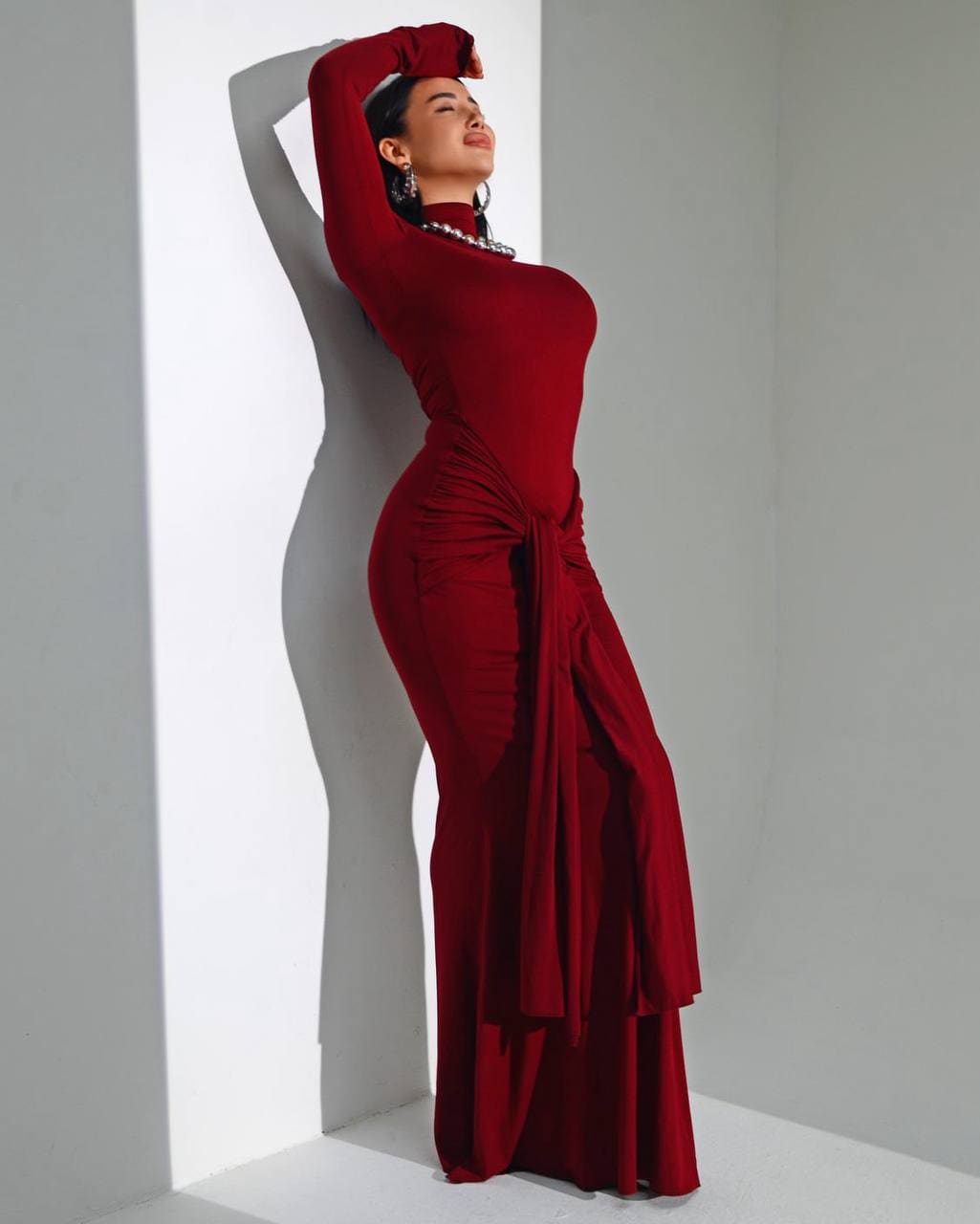 Profile shot of a woman in a burgundy dress against a white wall, arms raised above her head in a gathered waist gown.