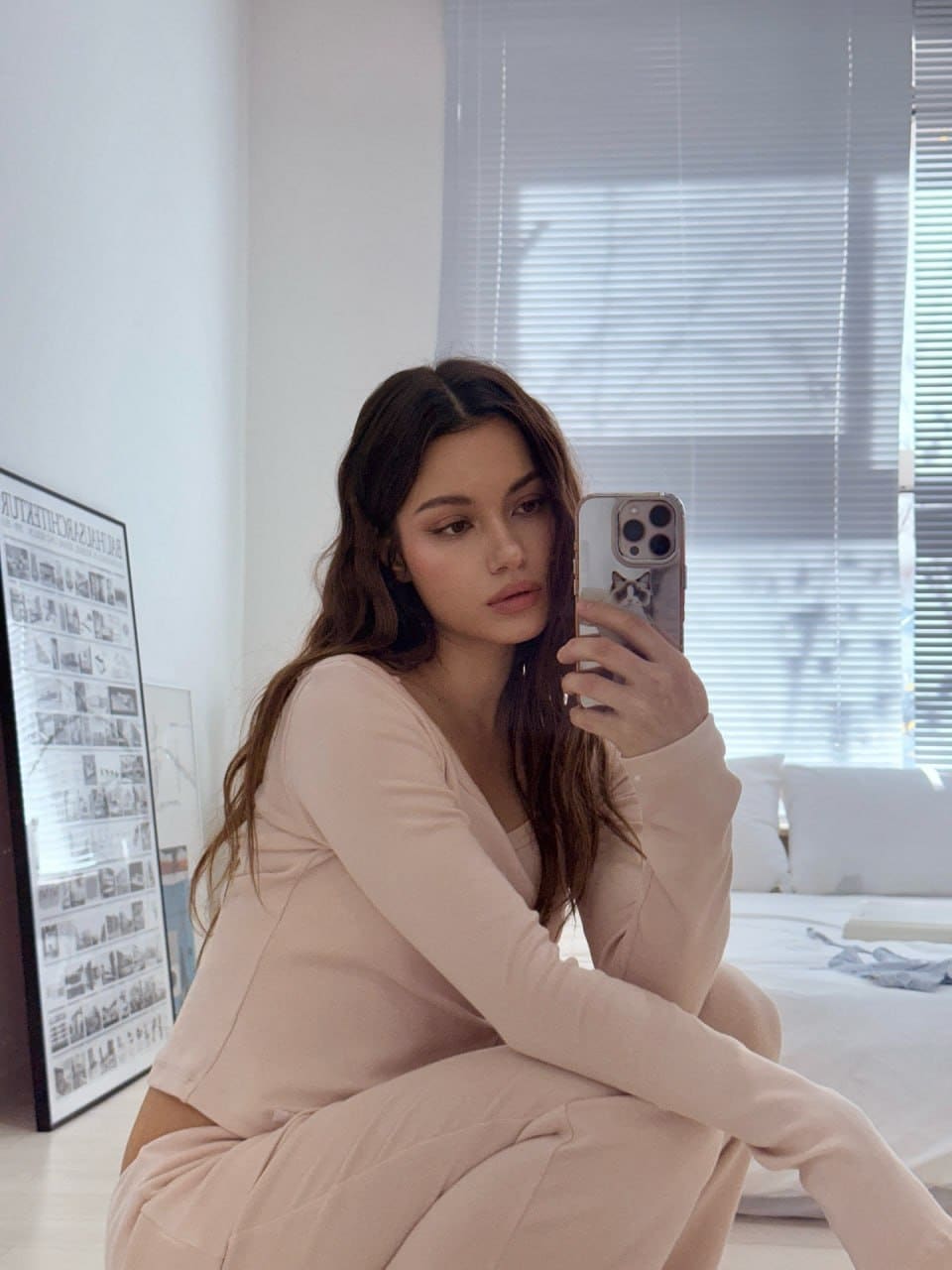 A mirror selfie shows her sitting on the floor in a matching light pink long-sleeved set, with a bed and framed poster behind her.