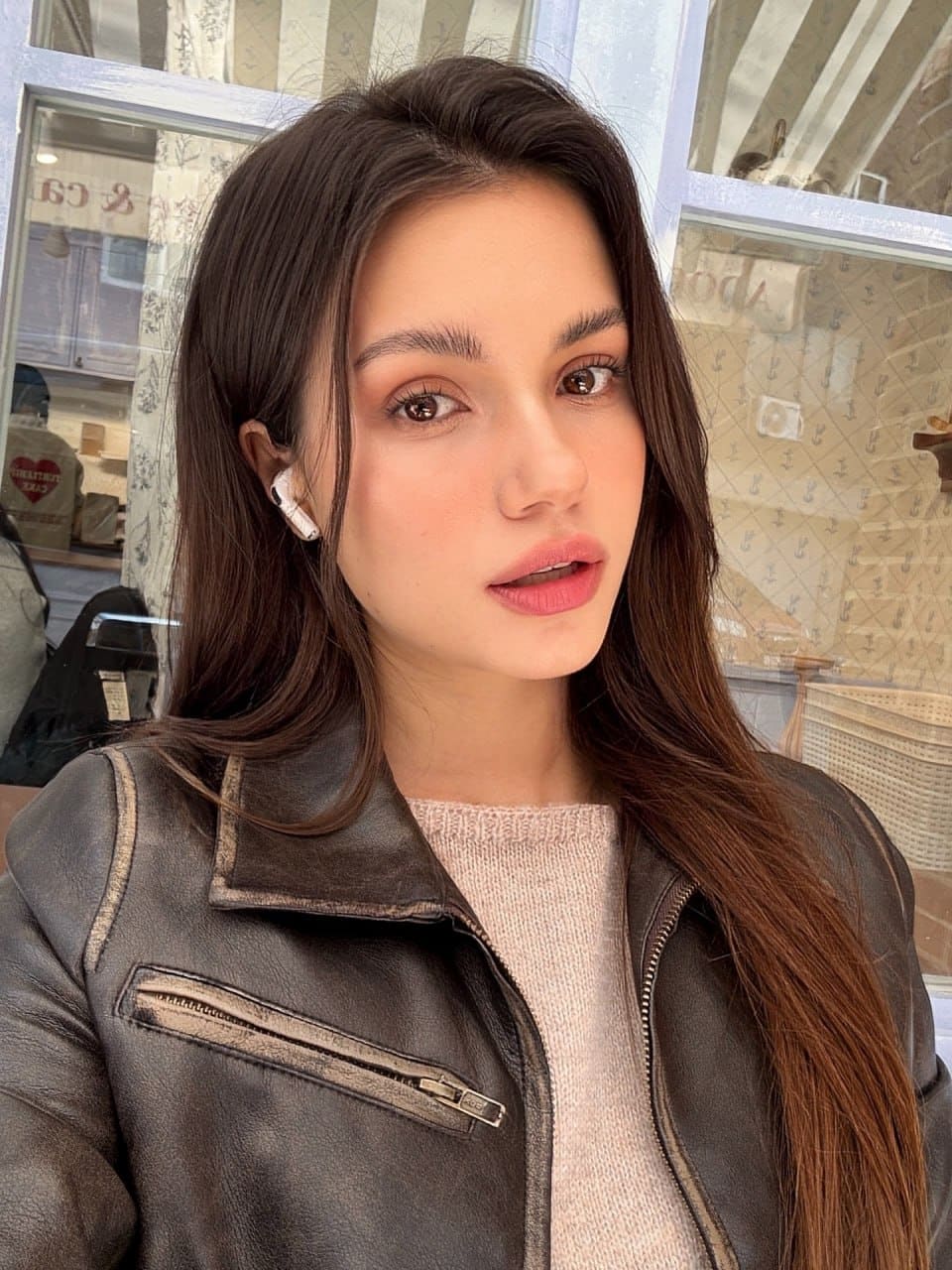 Mira takes a selfie in a dark leather biker jacket and pink knit sweater, wearing a white earbud, before a windowed storefront.