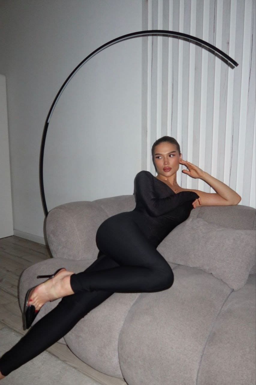 Reclining on a grey sofa, Daisy extends one leg while wearing a long-sleeved black bodysuit and pointed heels.