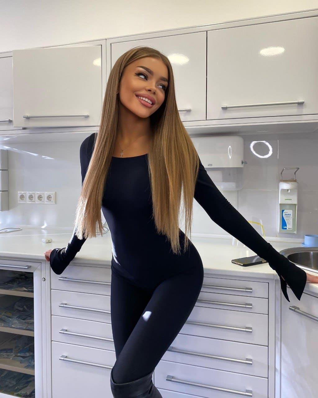 Halia leans back against a white kitchen counter in a black long-sleeved bodysuit with thumbholes, black leggings, and tall boots.