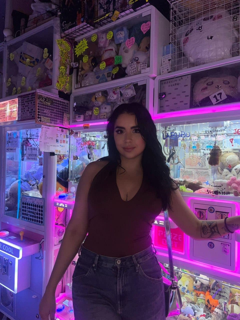 Rosa stands before illuminated claw machines in a maroon sleeveless top and blue jeans, showing an arm tattoo.