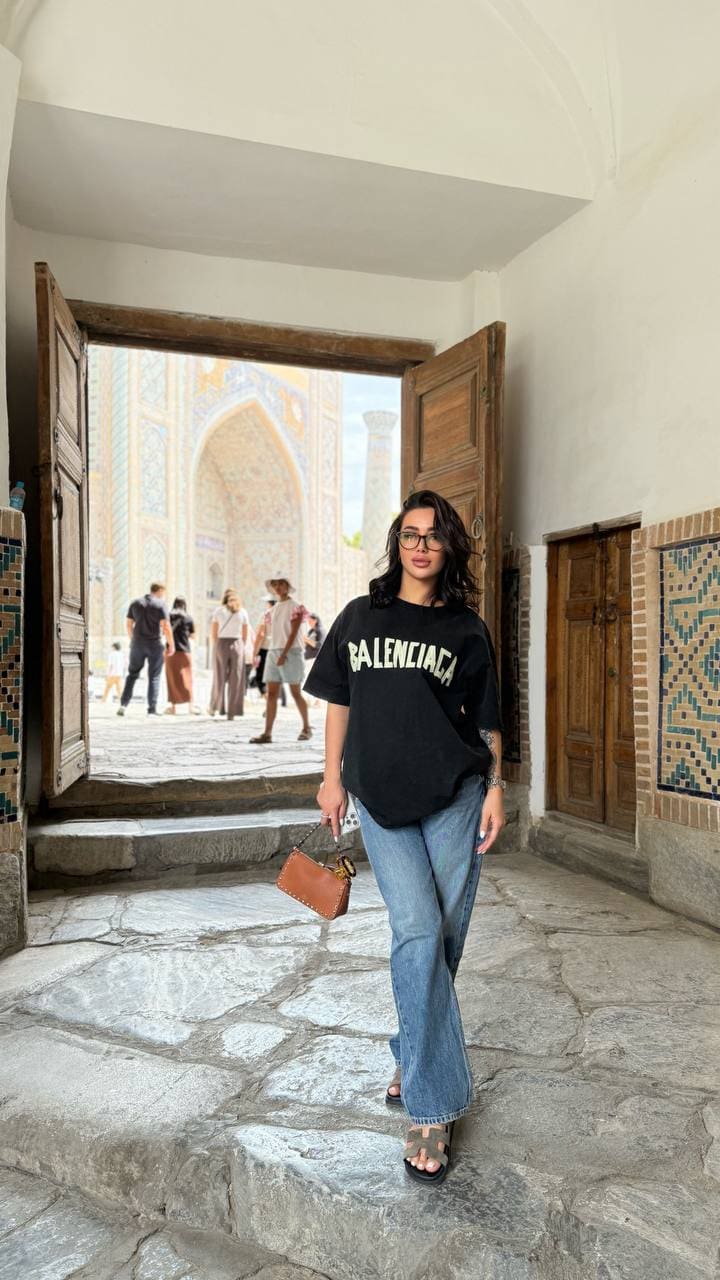 Standing on stone steps in a tiled entryway, a woman in an oversized black t-shirt, jeans, and sandals holds a brown bag.