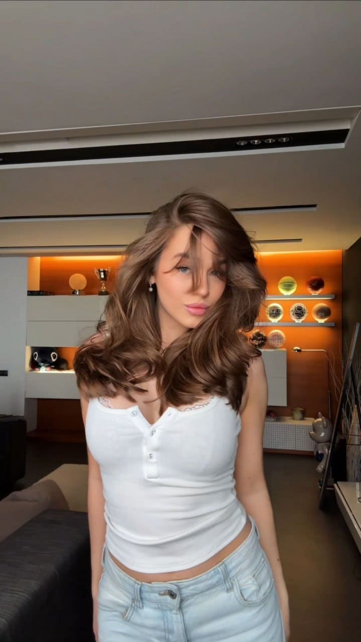 Amira tilts her head slightly while standing indoors in a white henley top and light blue jeans, illuminated by warm orange accent lighting.