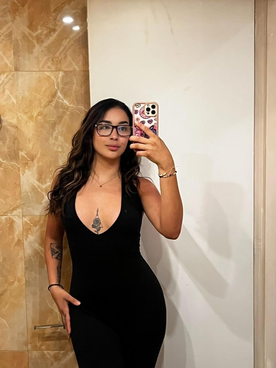 In a mirror selfie, Rosa wears glasses and a black bodysuit with a deep V-neck revealing a chest tattoo.