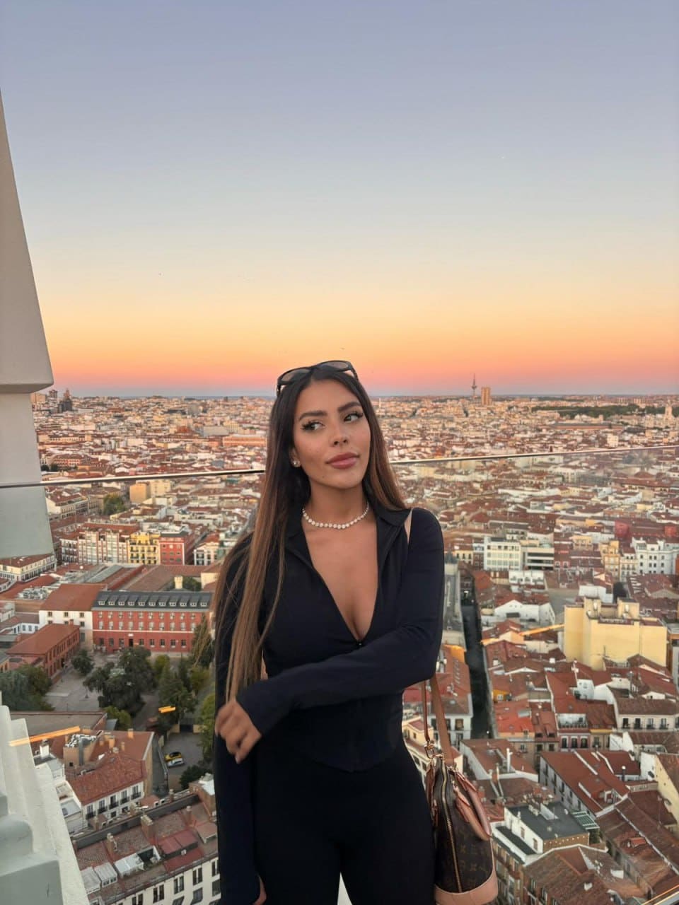 Alana stands on a rooftop terrace at sunset wearing a black long-sleeved bodysuit with a plunging neckline, holding a handbag.