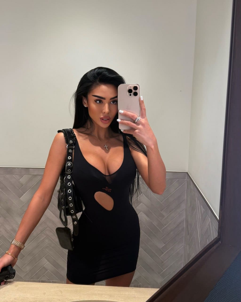 Inara captures a mirror selfie in a tight black mini dress with plunging neckline and mesh panel, accessorized with a studded bag.
