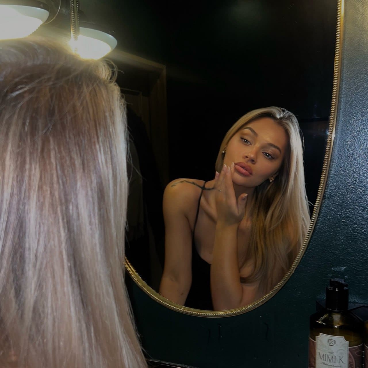 In a dimly lit room, Nora is captured in a mirror reflection wearing a black spaghetti-strap top, with one hand posed near her face.